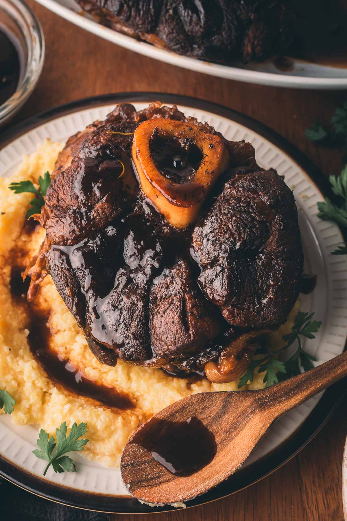 Braised osso buco with bone marrow served on creamy polenta, garnished with fresh parsley, and drizzled with sauce on a white plate.
