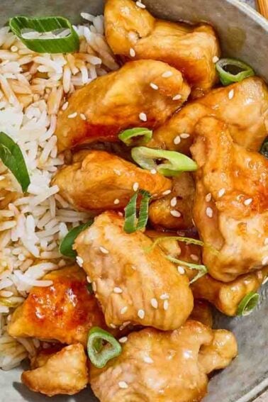 A bowl of white rice and glazed chicken pieces topped with sesame seeds and sliced green onions, with chopsticks and a striped napkin nearby—perfect for those seeking delicious budget chicken recipes.