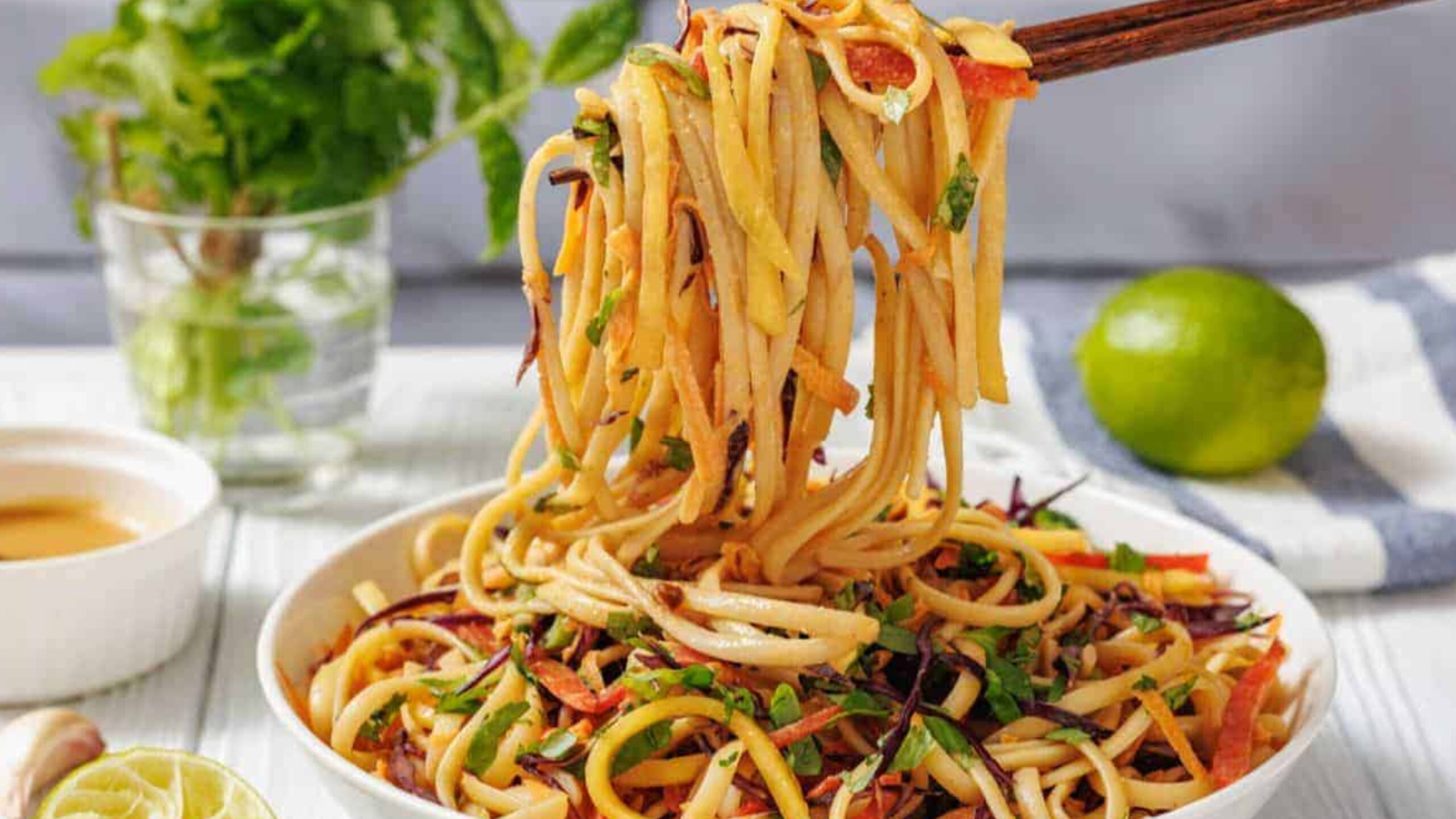 A bowl of Thai Noodle Salad with Mango and Spicy Peanut Dressing is lifted by chopsticks.
