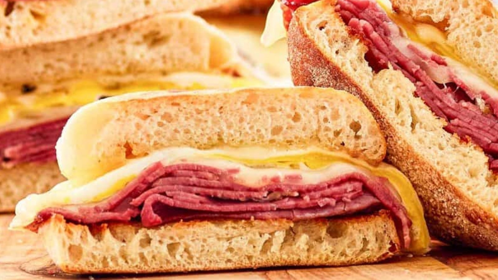 Close-up of sliced sandwich halves filled with layered sliced meat and melted cheese between pieces of bread on a wooden surface.