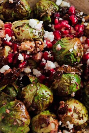 A bowl of roasted Brussels sprouts topped with crumbled feta cheese, pomegranate seeds, and chopped walnuts, with a spoon on the side.