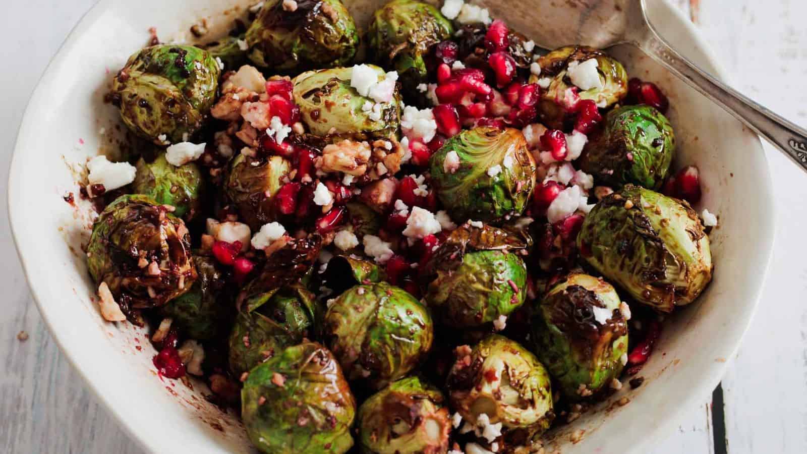 A bowl of roasted Brussels sprouts topped with crumbled feta cheese, pomegranate seeds, and chopped walnuts, with a spoon on the side.