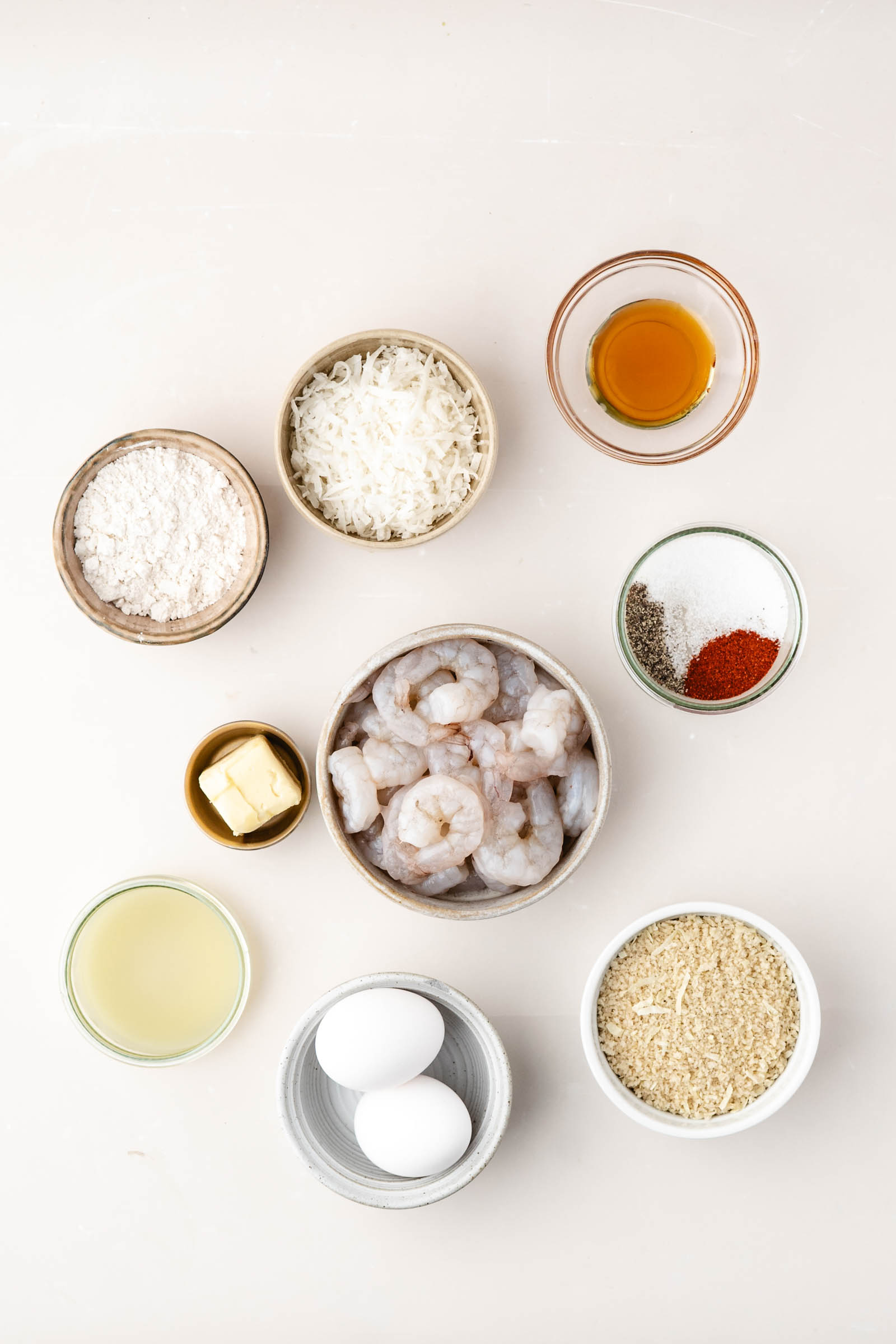 Overhead view of ingredients for coconut shrimp dish, including raw shrimp, flour, shredded coconut, eggs, spices, breadcrumbs, butter, oil, and a small bowl of liquid.