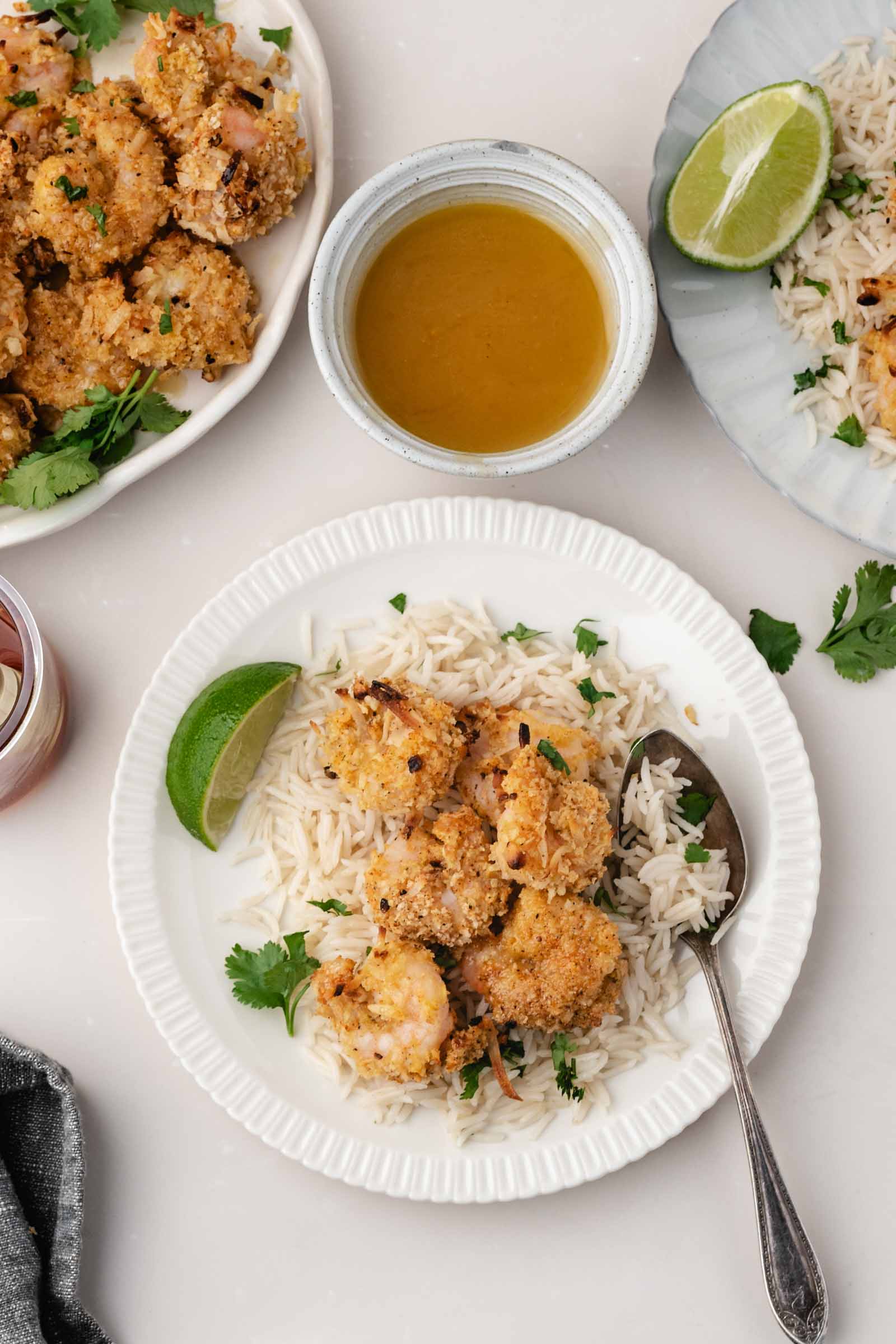 A white plate with coconut shrimp on white rice, garnished with cilantro and a lime wedge, with sauce and more shrimp on the side.