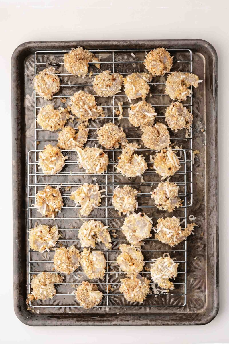 A baking sheet with a cooling rack holds several pieces of breaded coconut shrimp, spaced apart, prepared for baking.