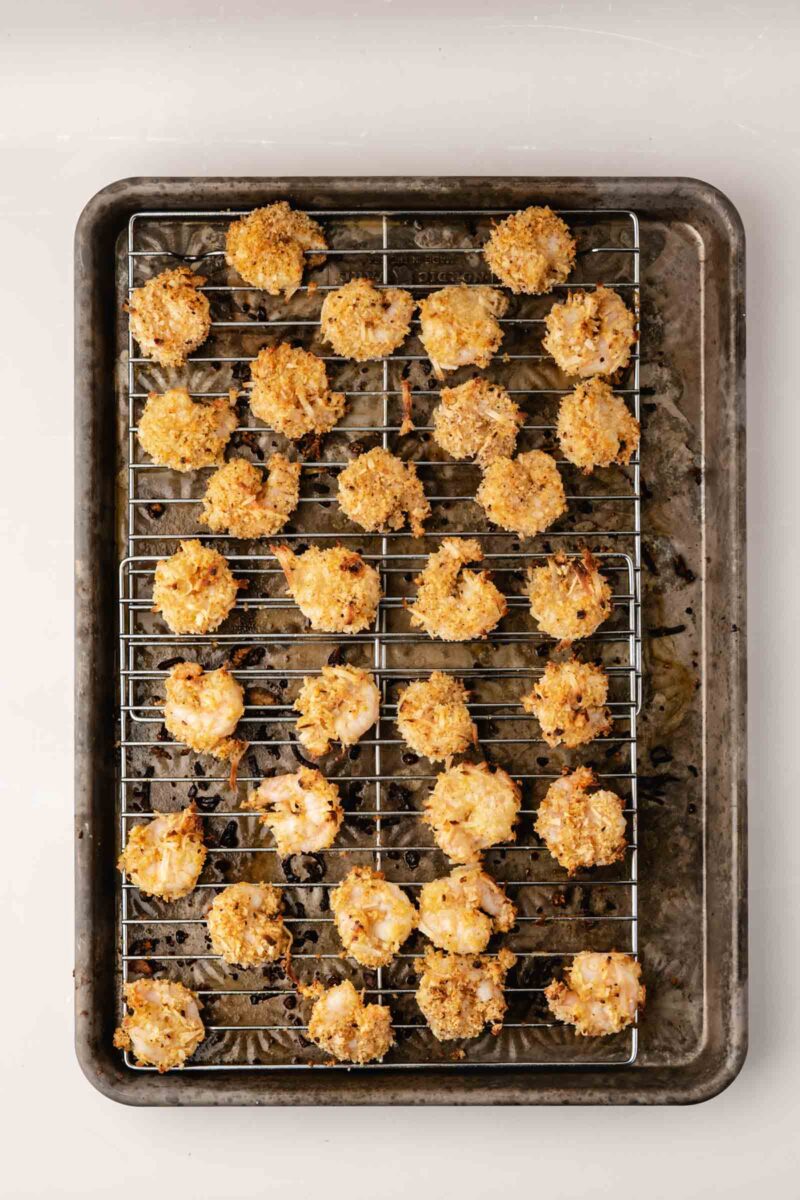 A baking sheet with a metal cooling rack holds baked coconut shrimp arranged in rows.