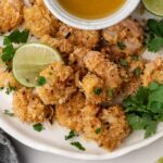 A plate of breaded coconut shrimp garnished with lime wedges and cilantro, served with a small bowl of dipping sauce.
