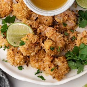 A plate of breaded coconut shrimp garnished with lime wedges and cilantro, served with a small bowl of dipping sauce.