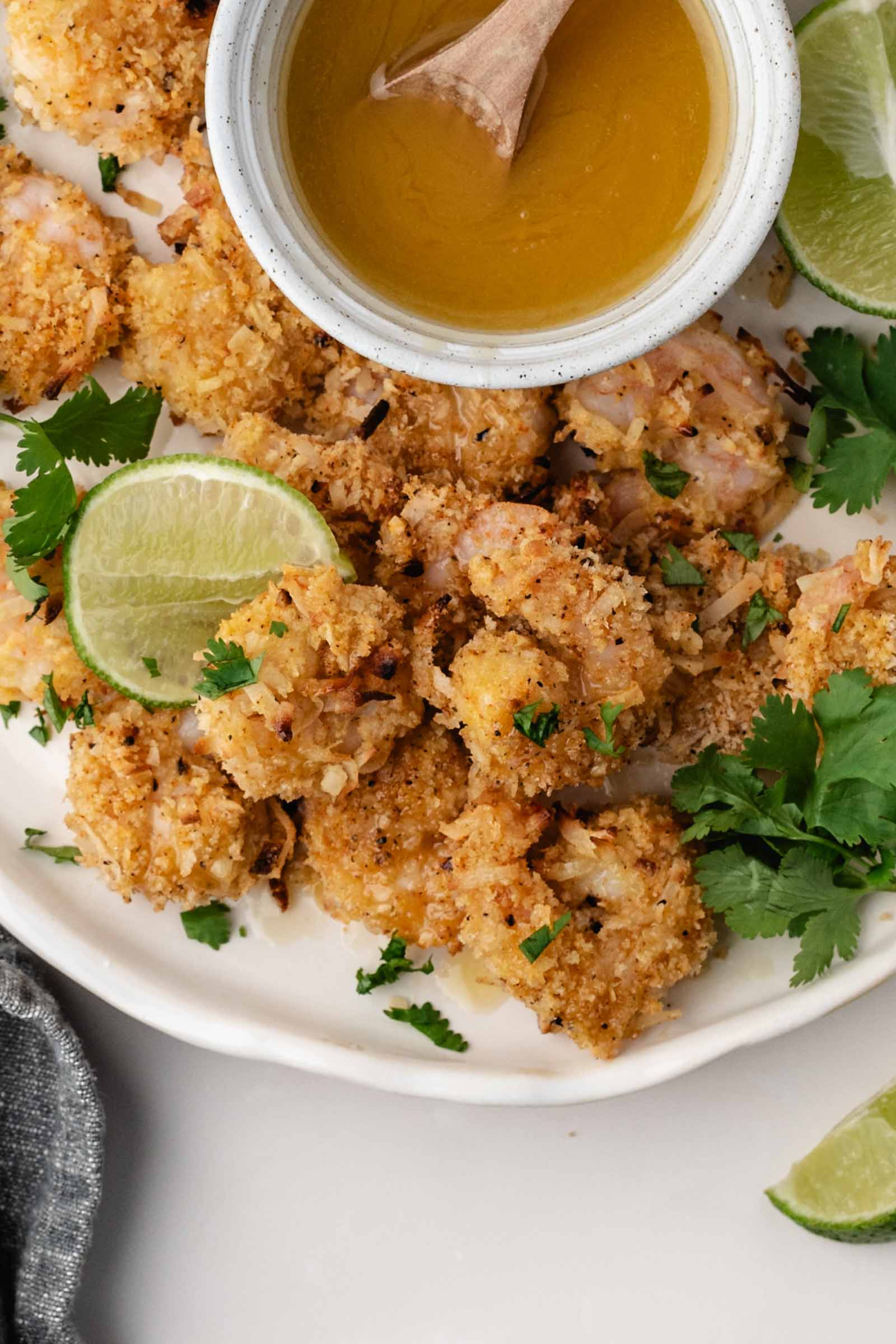 A plate of breaded coconut shrimp garnished with lime wedges and cilantro, served with a small bowl of dipping sauce.