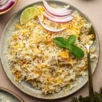 A plate of seasoned rice garnished with mint leaves, lime wedge, and sliced red onions, with a gold spoon on the side. Bowls of limes, onions, and yogurt are nearby.