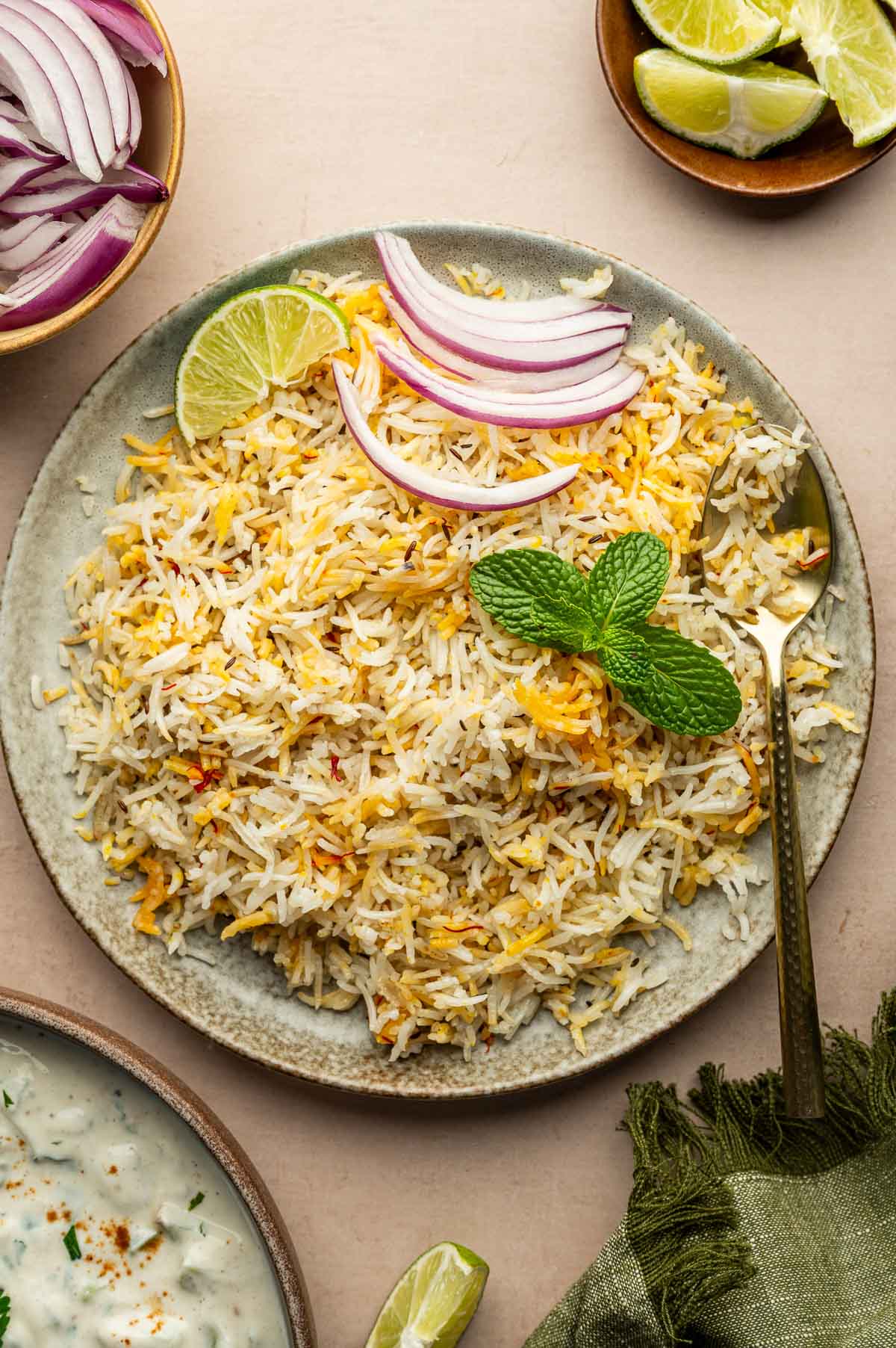 A plate of seasoned rice garnished with mint leaves, lime wedge, and sliced red onions, with a gold spoon on the side. Bowls of limes, onions, and yogurt are nearby.