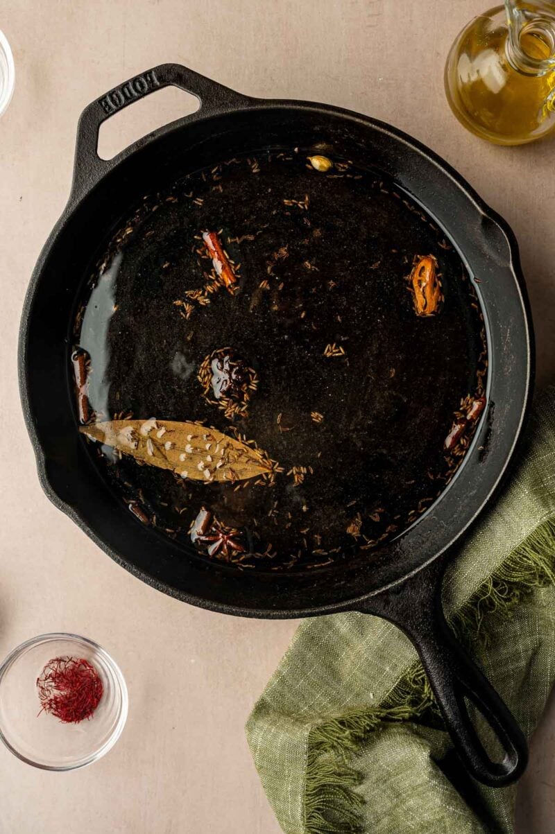 A black cast iron skillet containing hot oil with whole spices such as bay leaf, dried red chili, cumin seeds, and star anise, on a beige surface with a green cloth nearby.