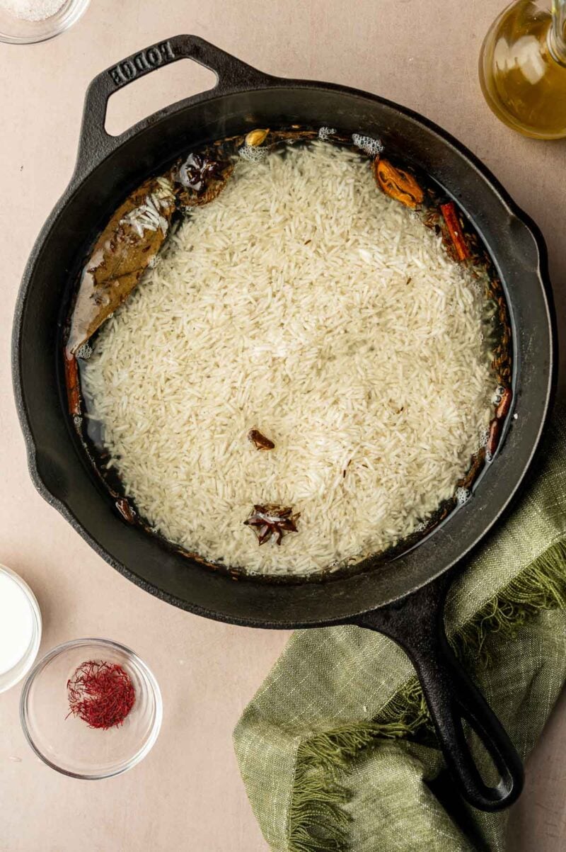 A cast iron skillet filled with uncooked rice, whole spices, and water sits on a countertop near a green cloth, a glass bowl of saffron, and a bottle of oil.