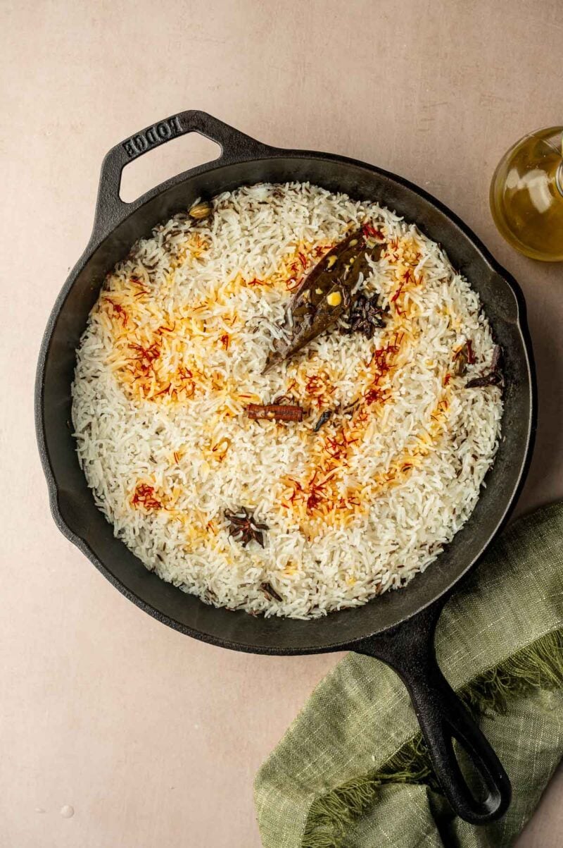 A cast iron skillet filled with cooked basmati rice, garnished with whole spices and saffron threads, sits on a light surface next to a green cloth and a bottle of oil.