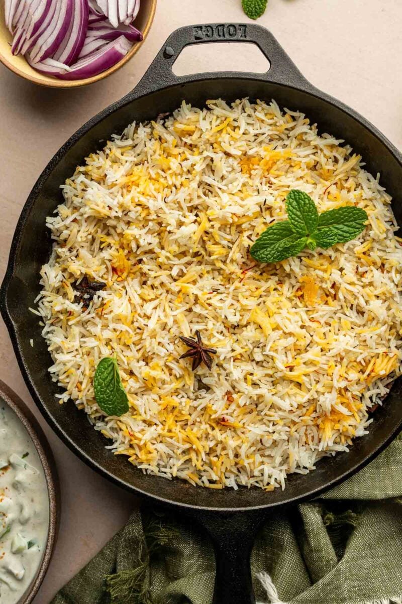 A cast iron skillet filled with saffron rice garnished with mint leaves and whole spices, surrounded by a bowl of sliced red onions and a green cloth.