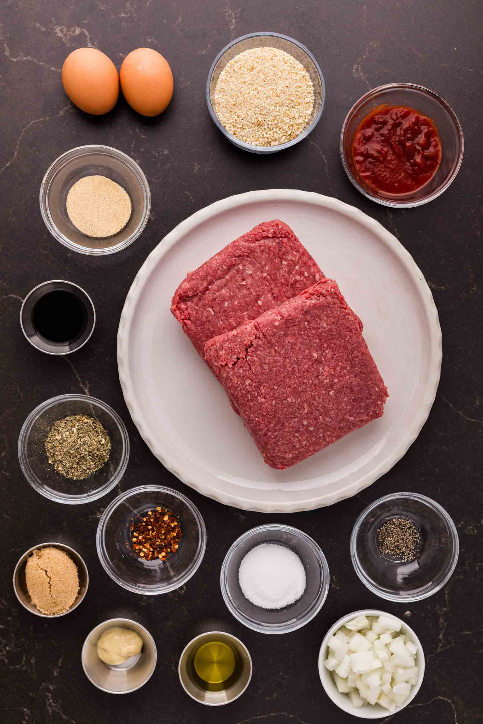Two blocks of raw ground bison on a plate, surrounded by bowls of eggs, breadcrumbs, ketchup, seasonings, chopped onion, and other meatloaf ingredients on a dark surface.