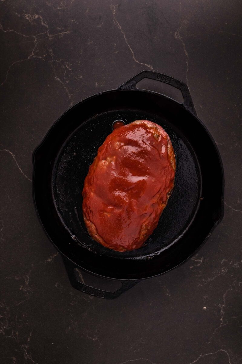 A cooked bison meatloaf covered in chili sauce sits in a black cast iron skillet on a dark, marbled surface.