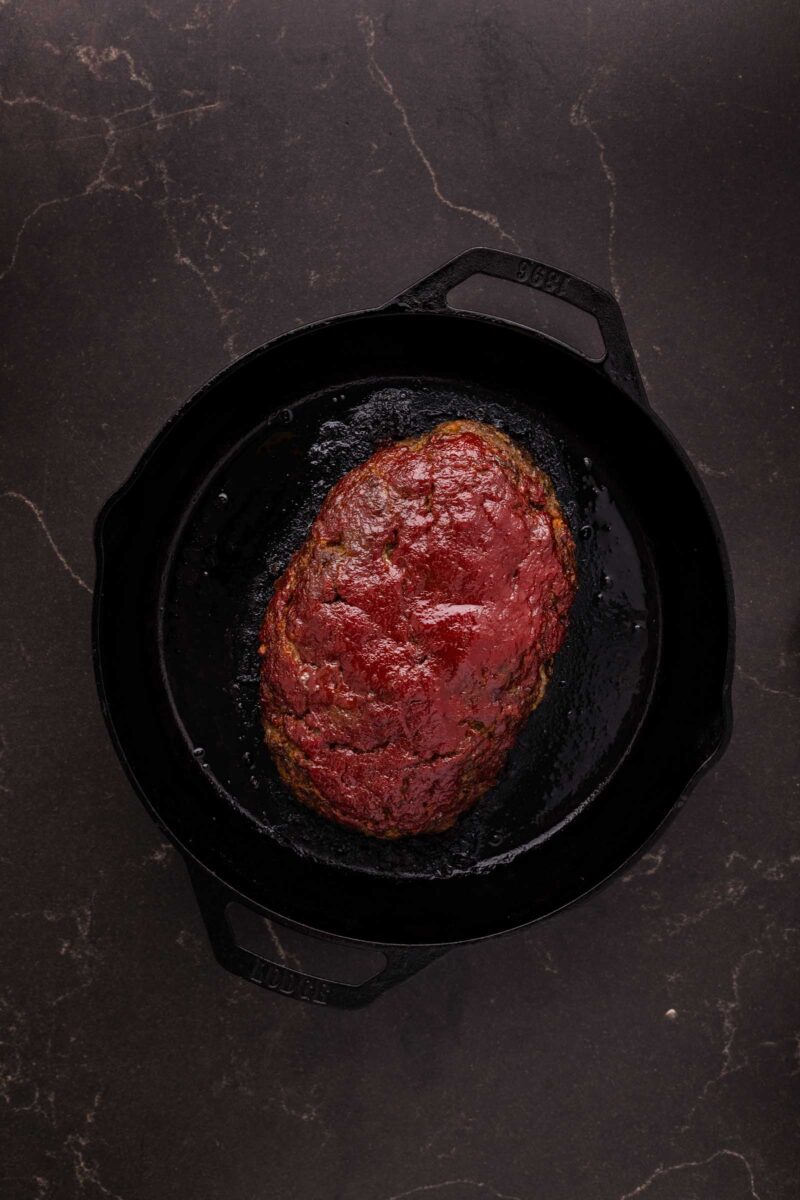 A cooked bison meatloaf with a chili glaze sits in a black cast iron skillet on a dark countertop.