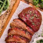 Sliced bison meatloaf with a ketchup glaze on parchment paper, garnished with chopped herbs, surrounded by bowls of green beans and fresh parsley.