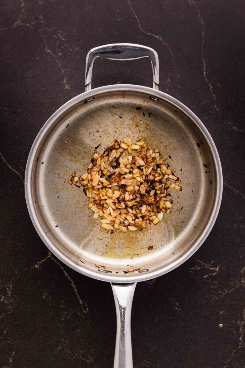 A stainless steel saucepan containing sautéed, browned onions sits on a dark countertop.