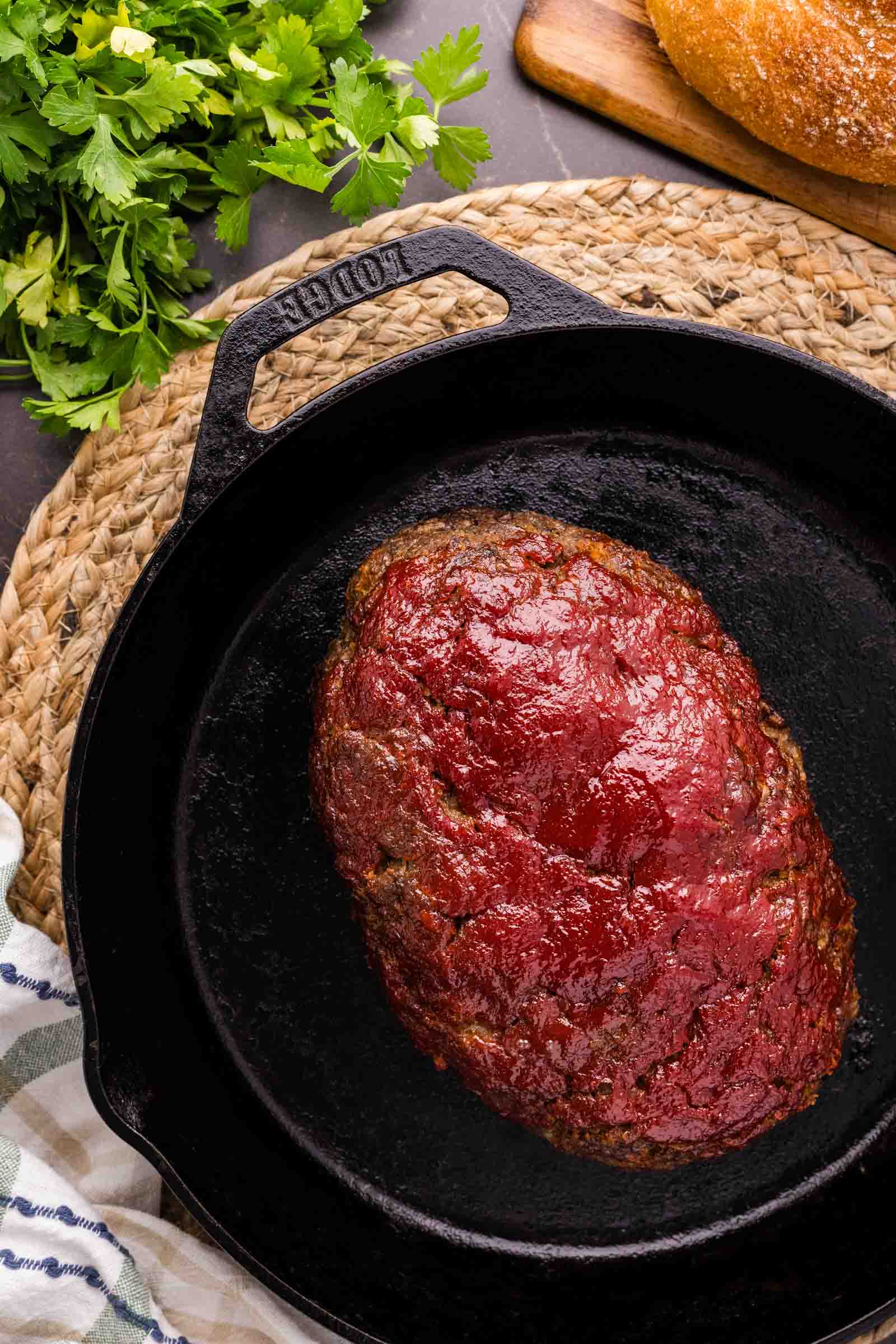 A cooked bison meatloaf with a tomato glaze sits in a cast iron skillet on a woven placemat, with fresh parsley and bread nearby.
