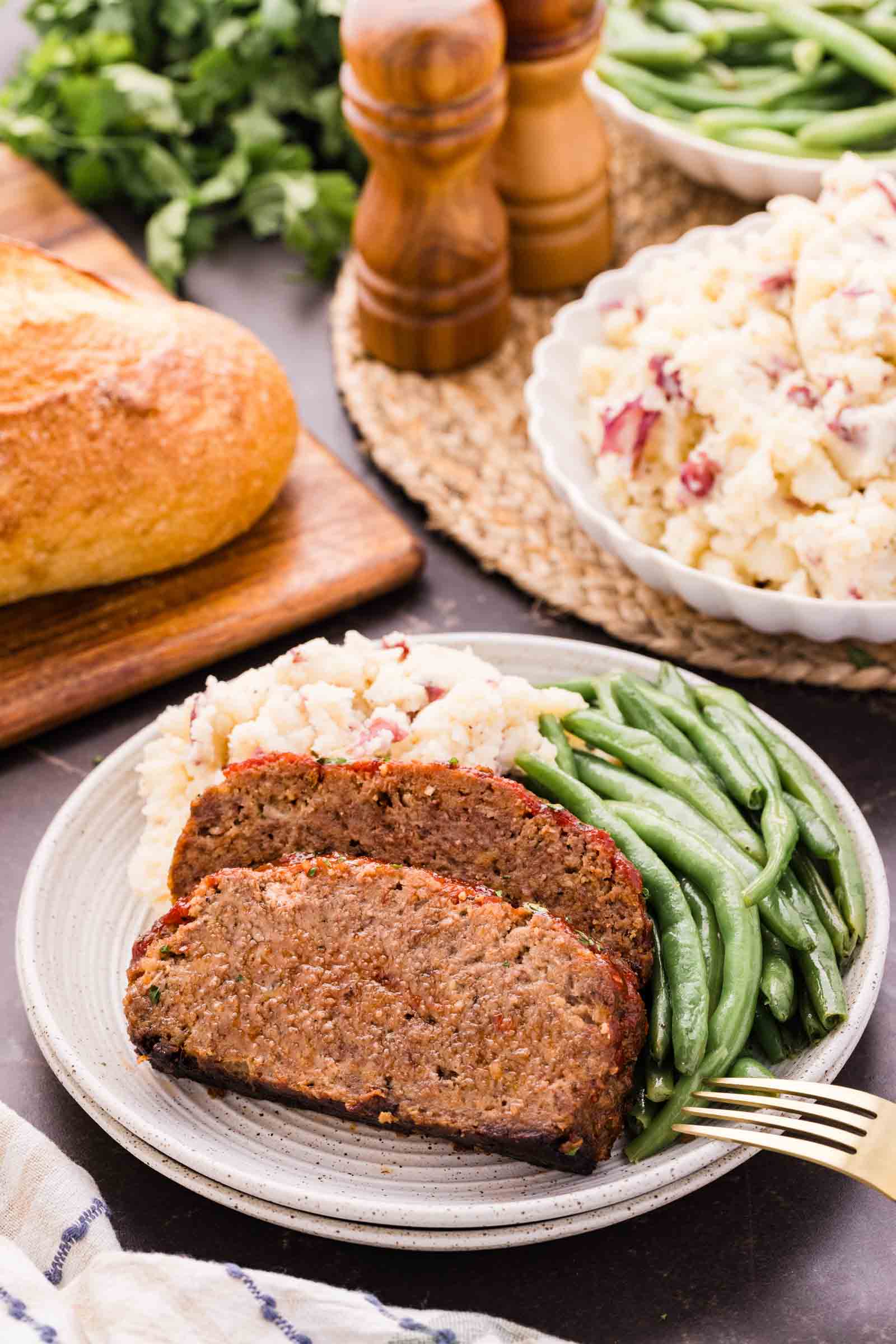 A plate with two slices of bison meatloaf, green beans, and mashed potatoes. Bread, green beans, and pepper mills are in the background.