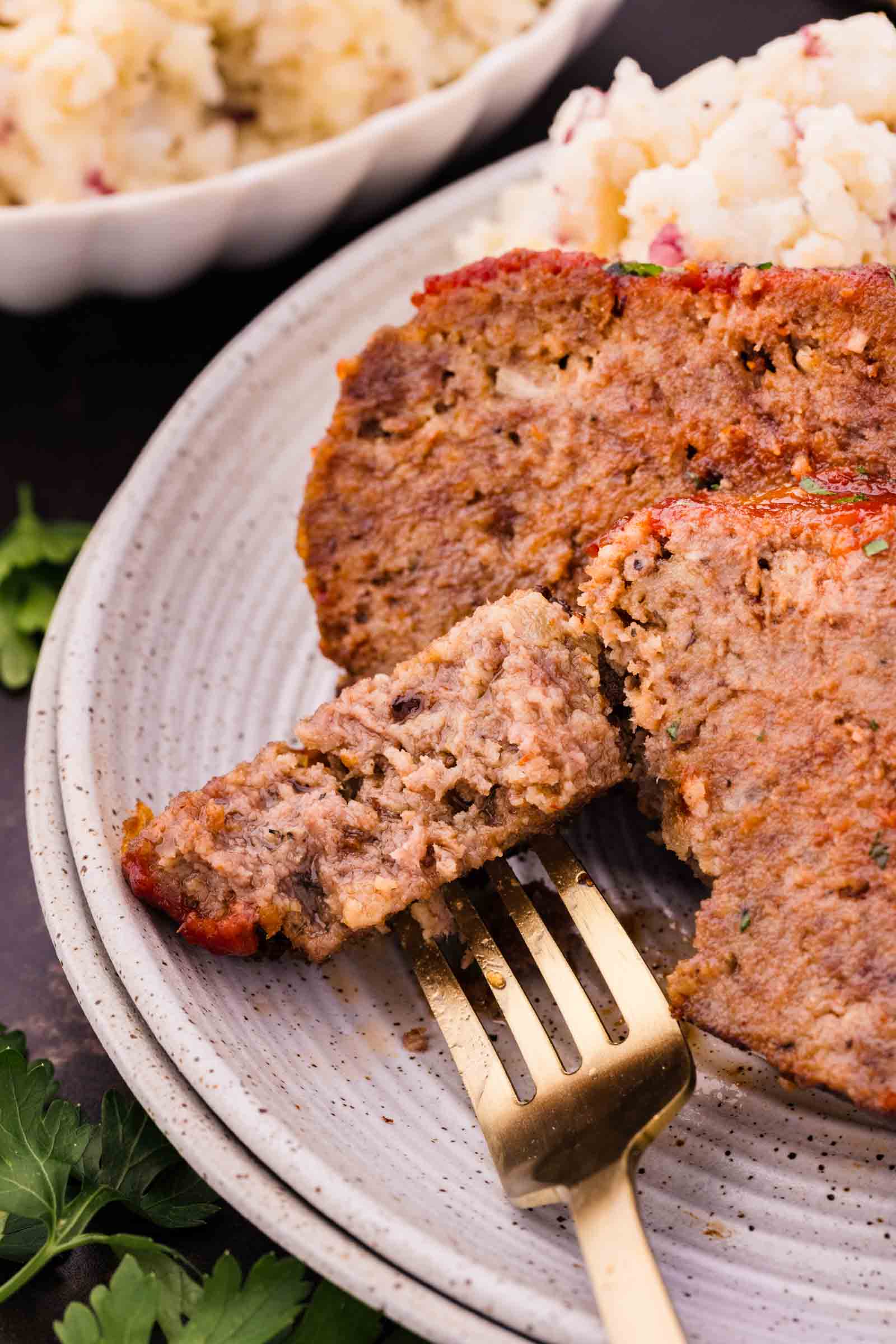 Close-up of sliced bison meatloaf on a plate with mashed potatoes in the background and a gold fork holding a piece of the meatloaf.
