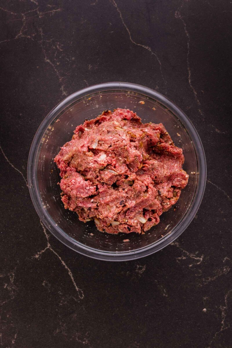 A clear glass bowl containing raw ground bison mixed with seasonings, placed on a dark countertop.