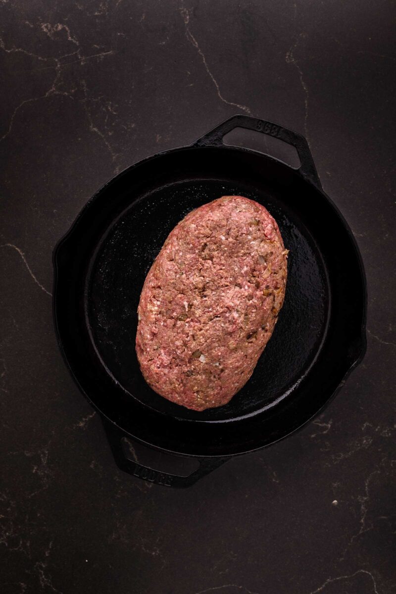 Raw bison meatloaf mixture shaped into a loaf sits in a black cast iron skillet on a dark, marbled surface.