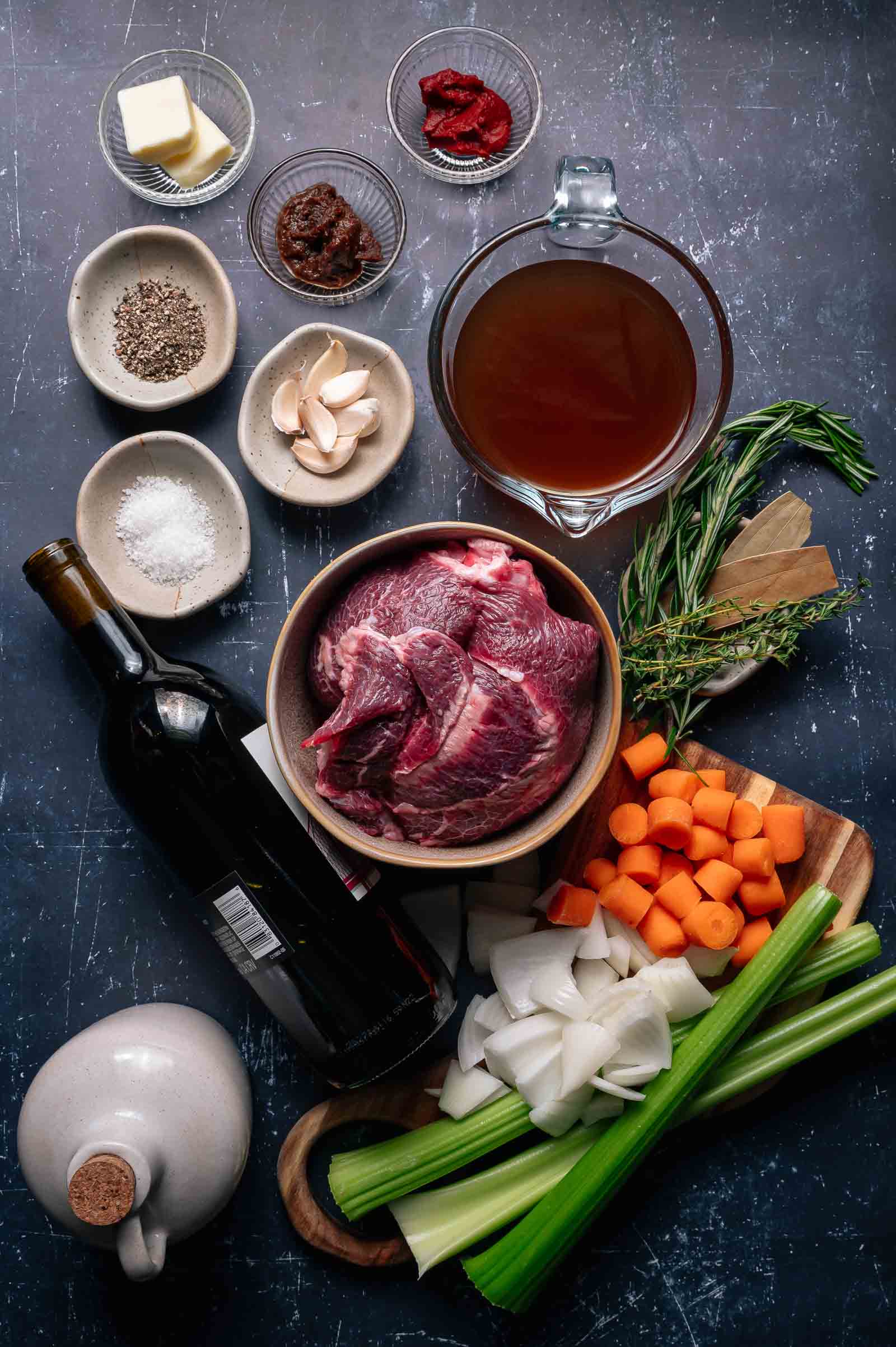Assorted ingredients for braised beef cheeks, including raw beef cheeks, carrots, celery, onion, garlic, herbs, butter, broth, wine, tomato paste, salt, pepper, and miso paste arranged on a dark surface.