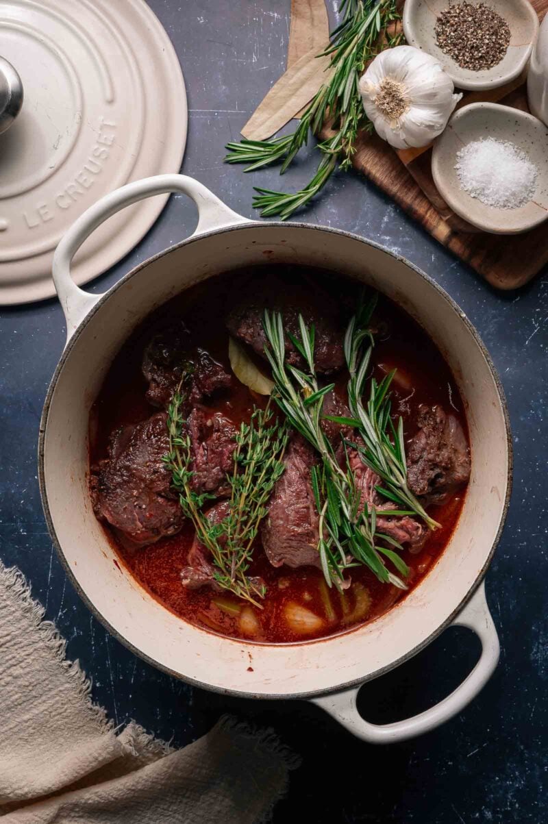 A white Dutch oven filled with beef cheeks, fresh rosemary and thyme and braising liquid sits on a dark surface next to a lid, garlic, spices, and herbs.