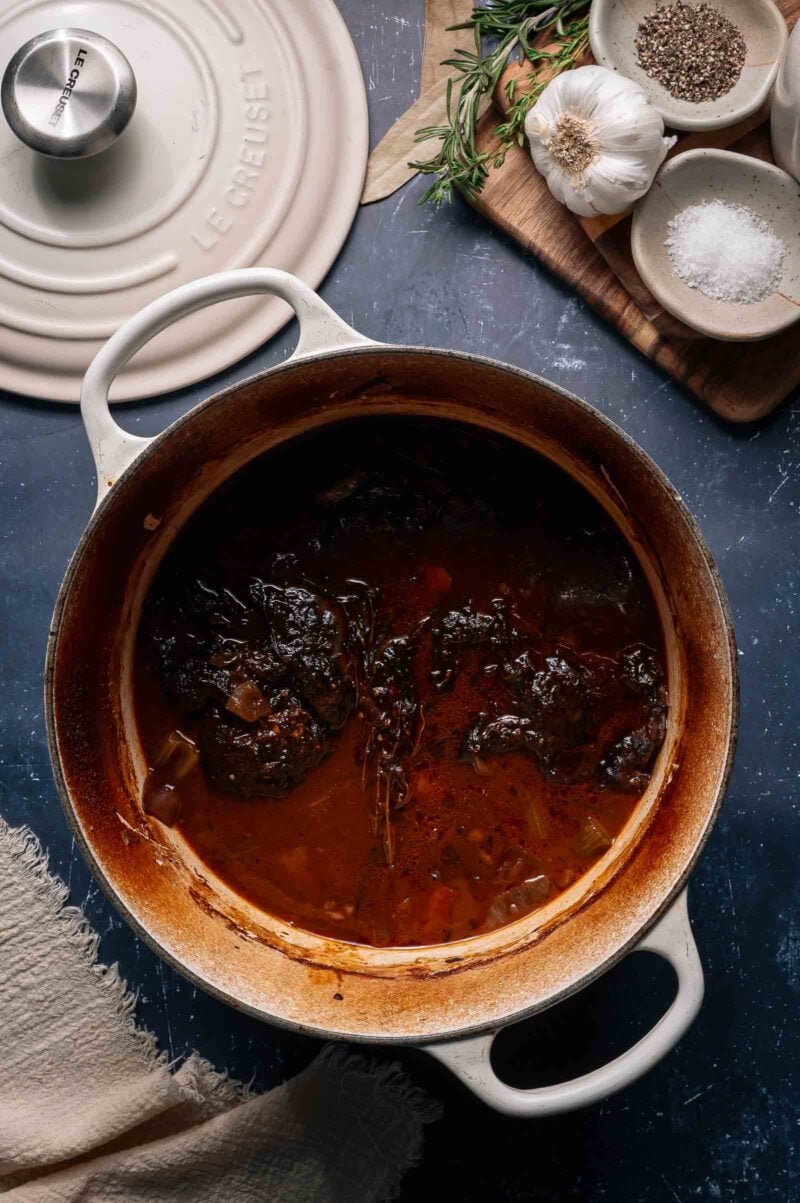 A white Dutch oven filled with braised beef cheels in a dark red wine sauce sits on a table beside a lid, herbs, garlic, salt, and pepper.