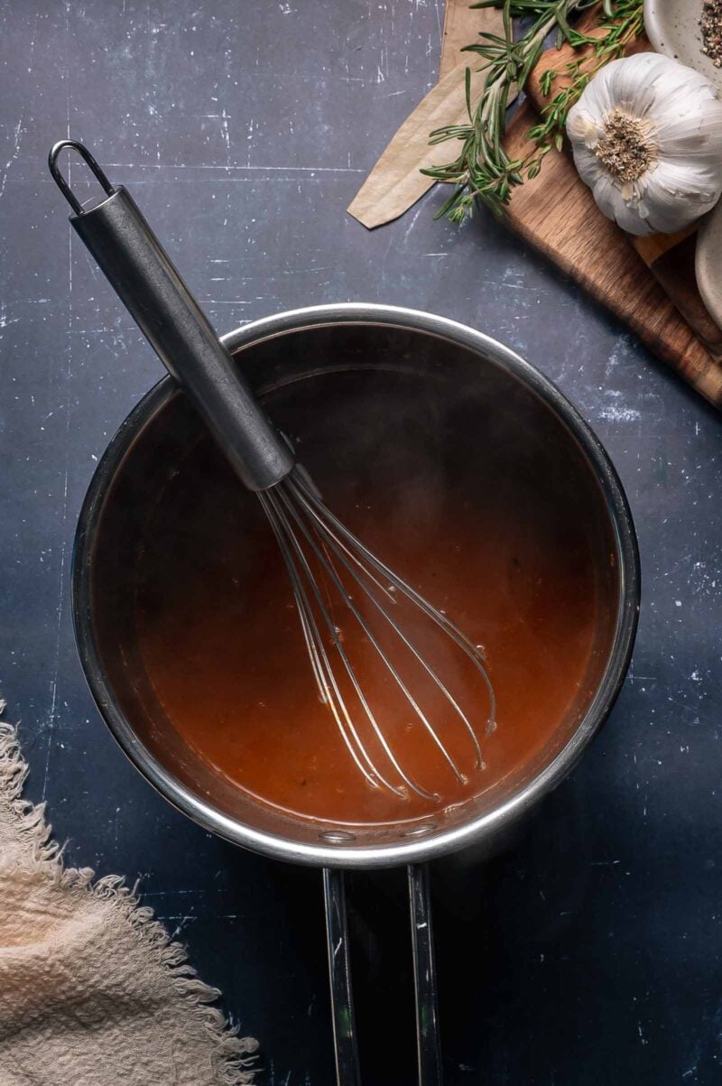 A metal whisk rests in a saucepan filled with the reduced braising liquid, with garlic, herbs, and a wooden cutting board nearby on a dark surface.