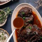 Oval plate with braised beef cheeks in a red qine sauce, garnished with chopped herbs. A ladle of sauce rests on the plate. Bowl of mashed potatoes and sprig of rosemary nearby.