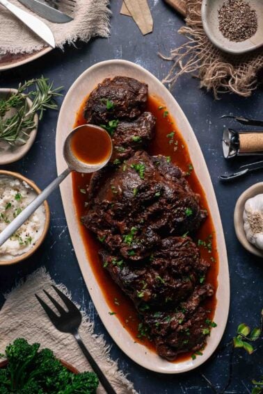 Oval platter with braised beef cheeks in rich red wine sauce, garnished with herbs, surrounded by mashed potatoes, spices, and fresh greens on a rustic table.