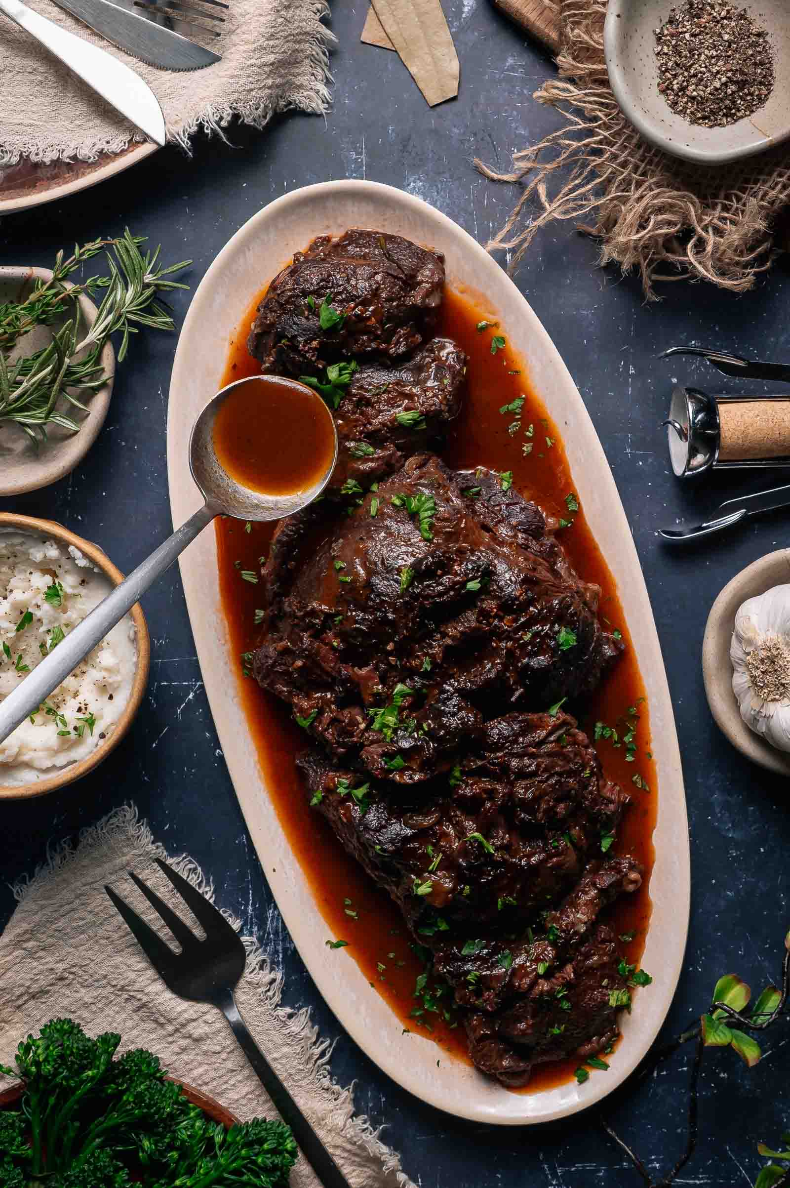 Oval platter with braised beef cheeks in rich red wine sauce, garnished with herbs, surrounded by mashed potatoes, spices, and fresh greens on a rustic table.