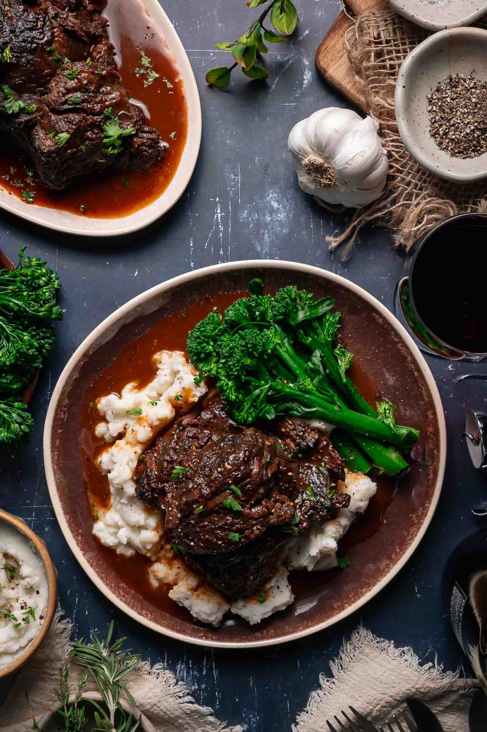 A plate of braised beef cheeks in sauce served over mashed potatoes with broccolini, surrounded by herbs, garlic, and seasonings on a dark table.