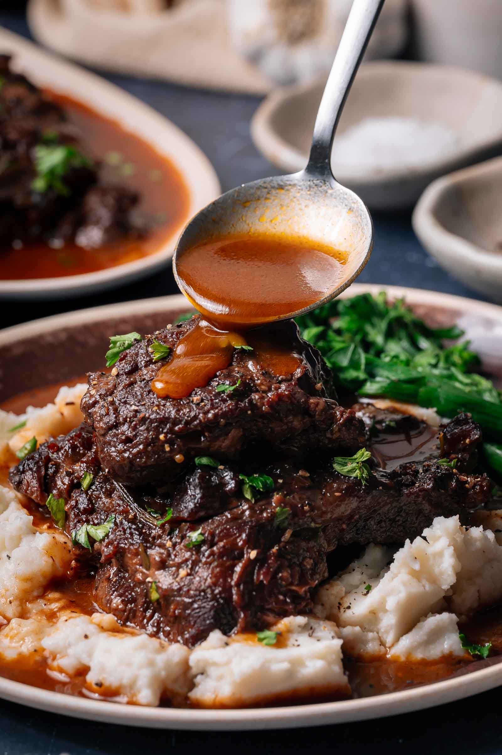 Braised beef cheeks served on mashed potatoes with a side of cooked greens, topped with sauce being poured from a ladle.