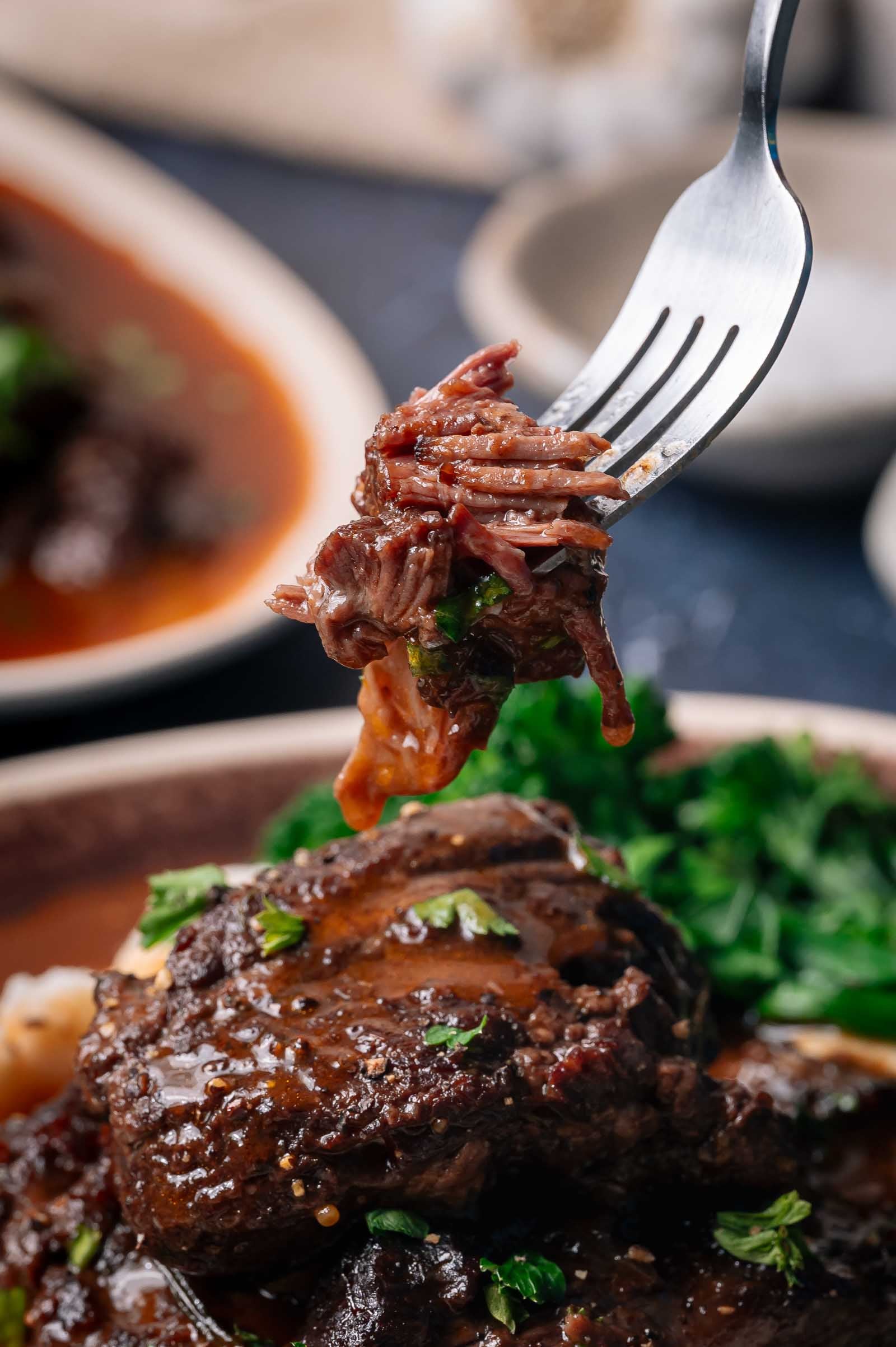 A fork holds a bite of tender, saucy beef cheeks above a plate with more beef and a side of greens.