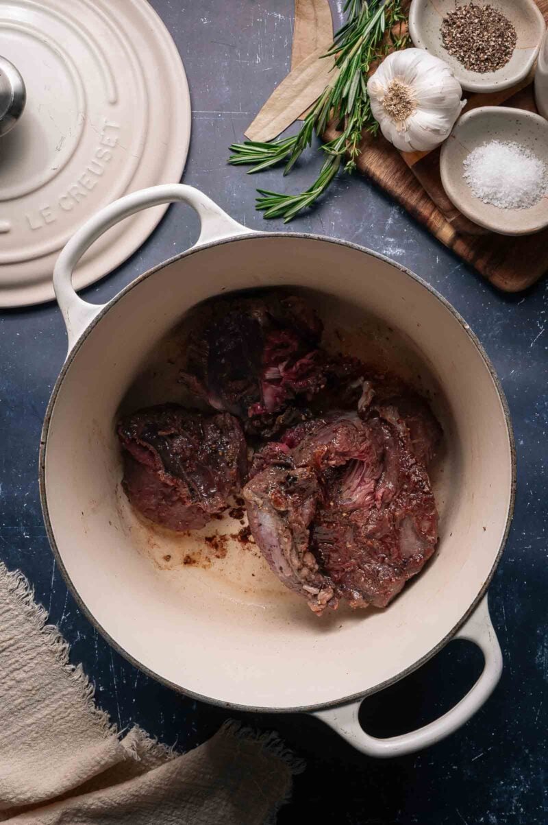 Beef cheeks searing in a white Dutch oven, surrounded by seasonings, fresh rosemary, garlic, and a linen towel.