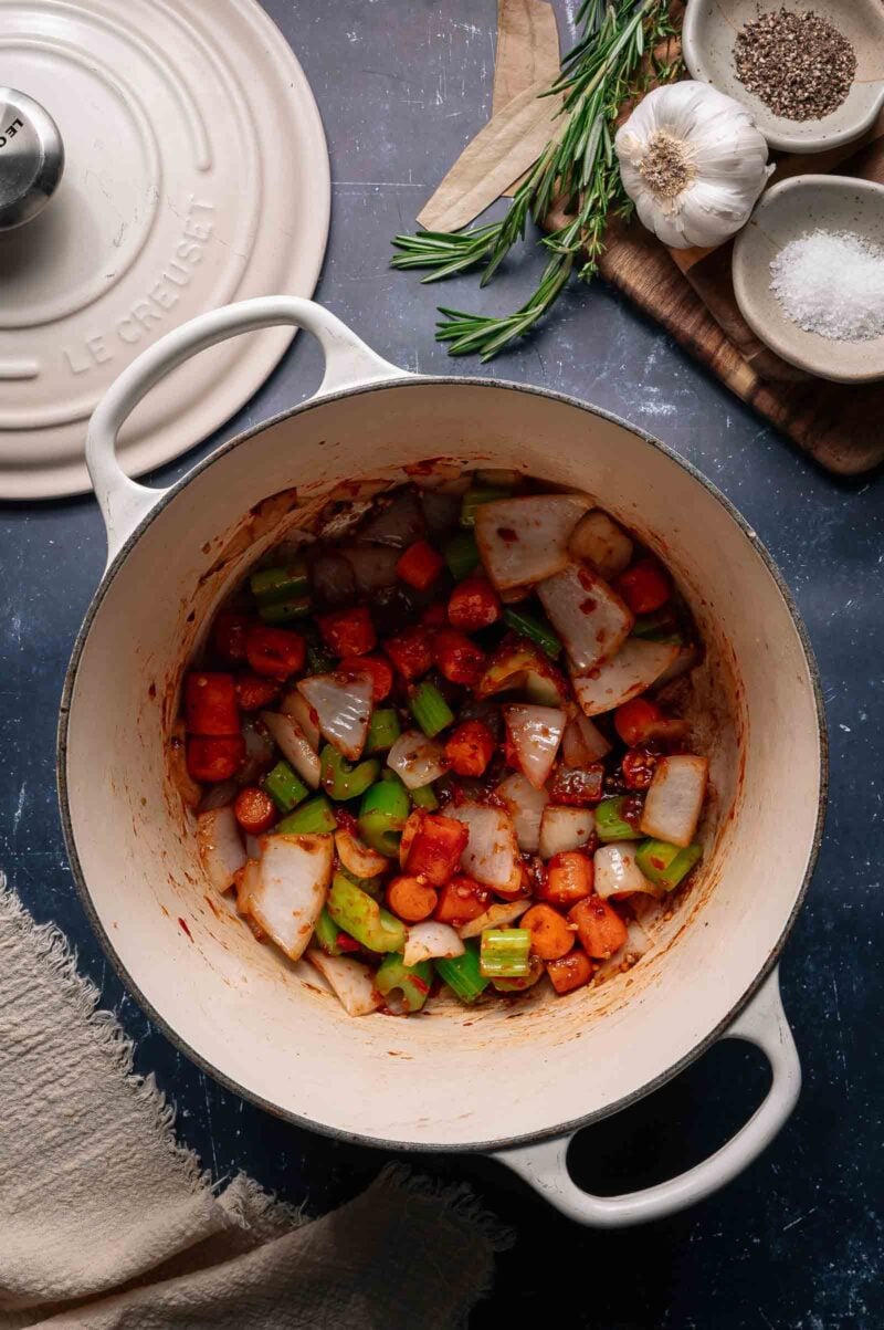 A Dutch oven with chopped onions, carrots, and celery being sautéed, surrounded by herbs, garlic, salt, and pepper on a dark countertop.
