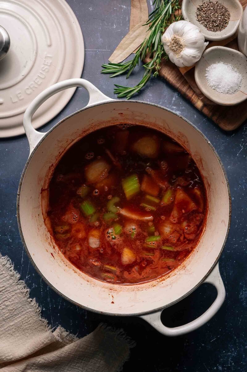 A white pot filled with vegetable soup sits on a dark surface; nearby are a lid, fresh herbs, garlic, salt, pepper, and a wooden board.