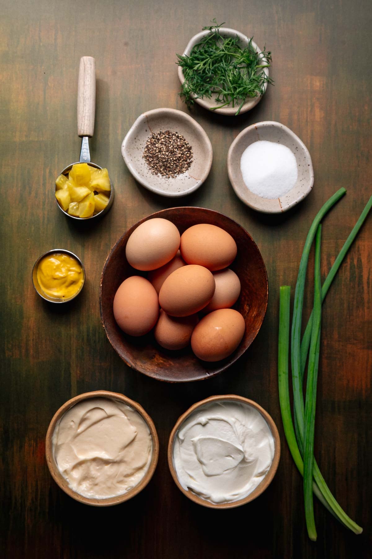 A variety of ingredients on a table, including eggs, chopped pineapple, mayonnaise, sour cream, fresh herbs, black pepper, salt, and whole green onions.