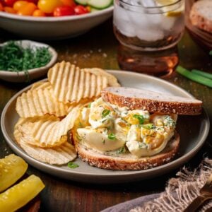 A plate with a broken egg salad sandwich on whole grain bread, potato chips, sliced pickles, and a drink with ice and lemon in the background.
