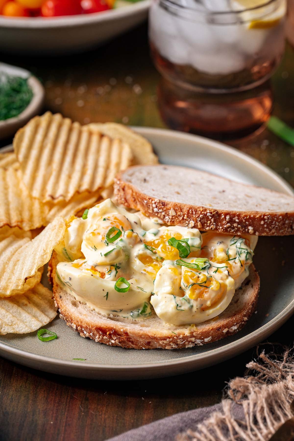 Open-faced creamy broken egg salad sandwich on multigrain bread served with potato chips on a plate; glass of iced drink and bowl of vegetables in the background.