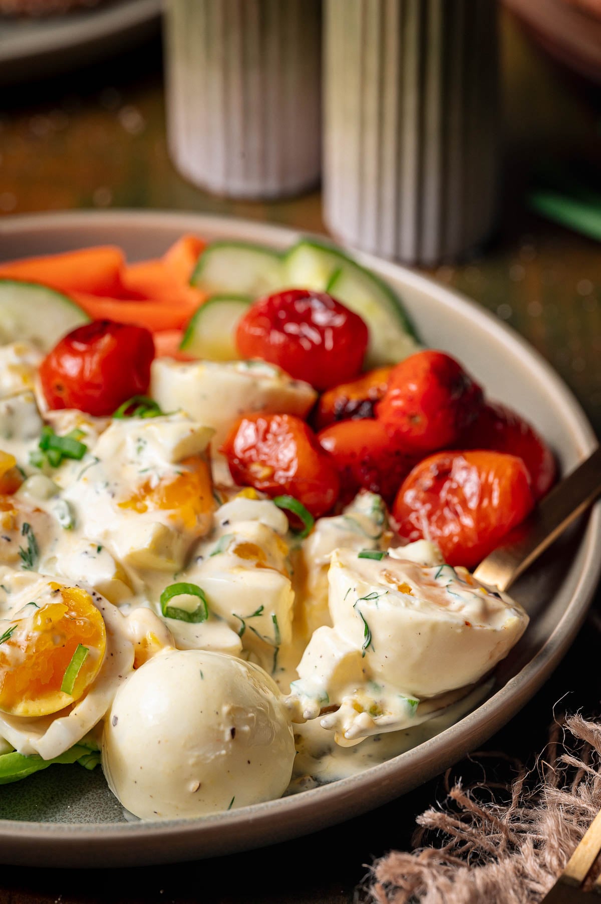 A plate with broken eggs in creamy sauce, blistered cherry tomatoes, sliced cucumber, and carrot sticks, garnished with chopped herbs.