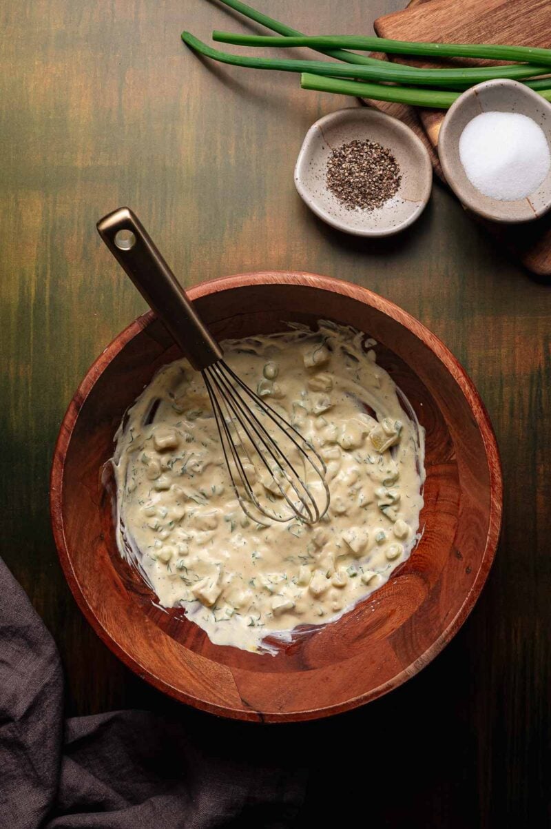 A wooden bowl contains creamy dressing with chopped herbs and a whisk; green onions, salt, and pepper are on the table nearby.
