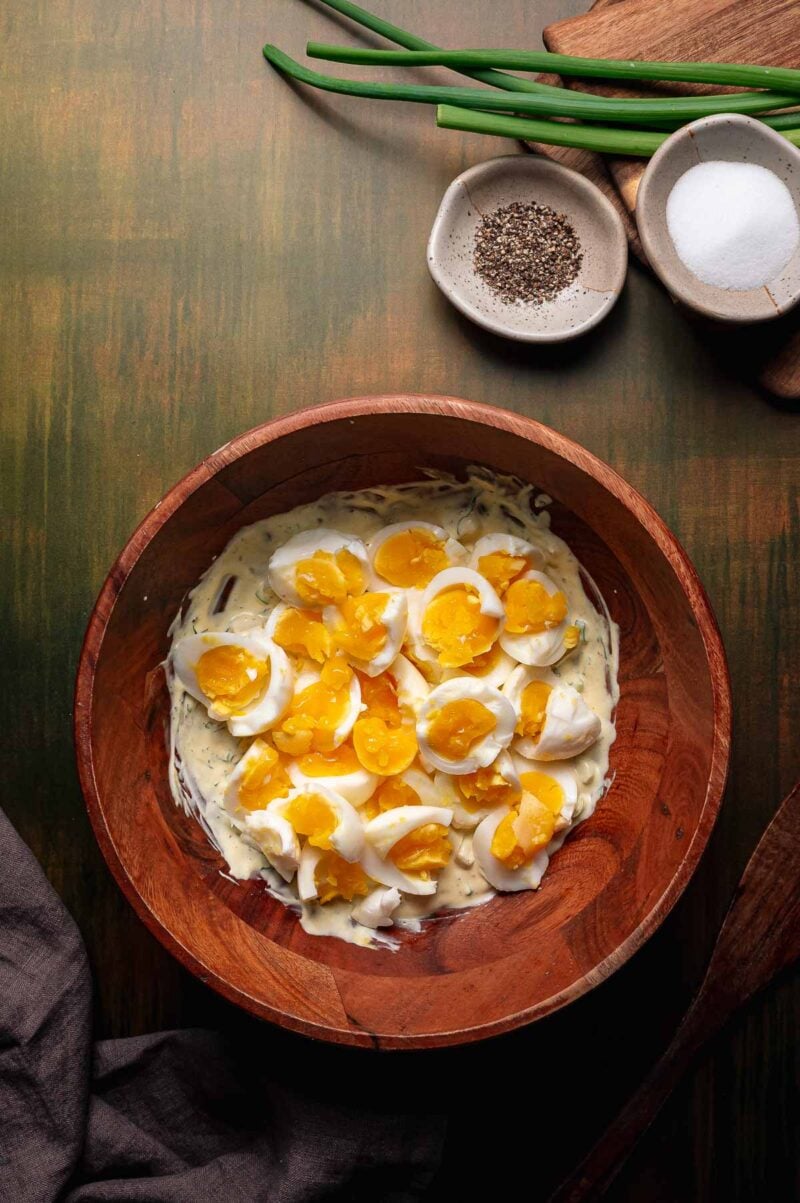 A wooden bowl with torn boiled eggs atop the dressing, next to bowls of salt, pepper, green onions, and a wooden cutting board on a green surface.