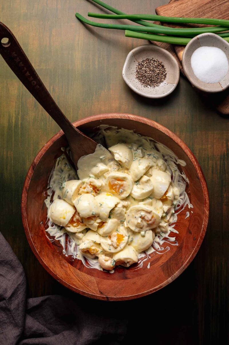 A wooden bowl filled with creamy broken egg salad being gentle folded with a spatula, with salt, pepper, green onions, and a napkin nearby on a wooden surface.