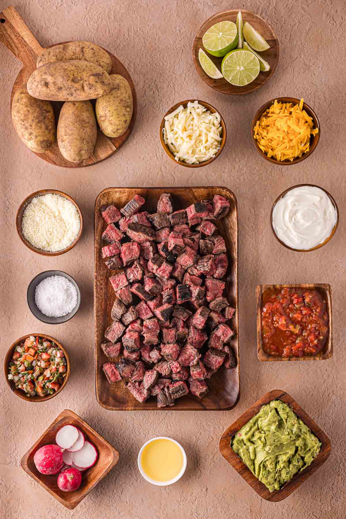 Top-down view of diced carne asada, potatoes, shredded cheese, lime wedges, guacamole, salsa, sour cream, pico de gallo, radishes, and other toppings arranged on a beige surface.